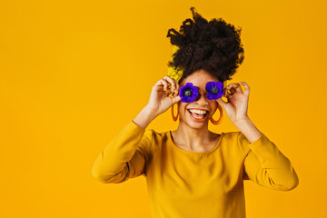 Portrait of a happy young woman holding two flowers over eyes, laughing and having fun, against yellow background - obrazy, fototapety, plakaty