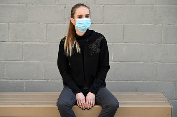 A model is sitting on a bench in a medical mask. Close-up portrait of a young blonde Caucasian woman in a city outdoors on a sunny day against the background of a gray wall of a building.