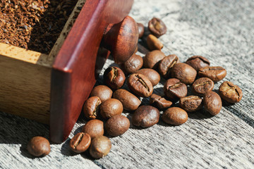 Roasted coffee beans are scattered on the table next to the red drawer of the coffee grinder and ground coffee. Photo in bright sunlight in the morning.