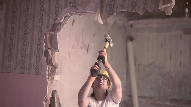 Slow motion video of a young hard working independent woman breaking a wall with a hammer to renovate her home wearing protective personal equipment for safety