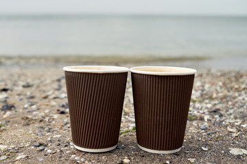 Two disposable paper cups of coffee standing on sand against blue sea, copy space. Take-out coffee