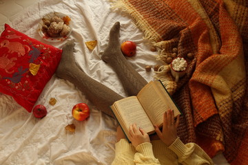 Top view of an open book, female legs and hands on a bed with apples, sweets, a pillow and a plaid around