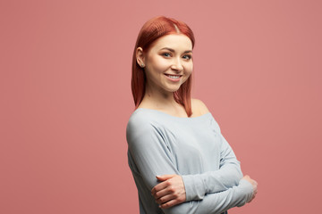 Obraz premium Smiling ginger girl in gray sweater with arms crossed standing in studio isolated on pink background