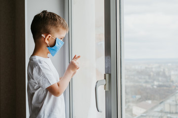 a child in a medical mask is sitting at home in quarantine because of coronavirus and covid -19 and looks out the window.