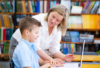 The teacher helps the boy to study on a laptop in the library. The boy looks at the laptop screen,...