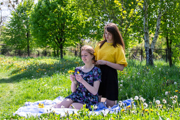 Young sisters in nature weave a wreath of dandelions. Talk and enjoy the rest. Weave flowers into the hair