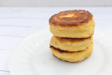 Three cheesecakes stacked on top of each other. Three fried curd cheesecakes on a white plate on a plank table. Delicious Breakfast of flour, eggs and cottage cheese