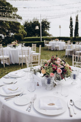 Wedding banquet. Tables in a green meadow are decorated with flower arrangements, on the tables are white tablecloths, plates with napkins, glasses and candles, cutlery. White bulbs hang over tables