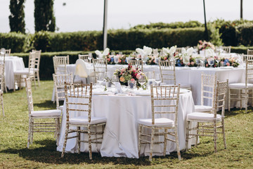 Wedding banquet. Tables in a green meadow are decorated with flower arrangements, on the tables are white tablecloths, plates with napkins, glasses and candles, cutlery. White bulbs hang over tables
