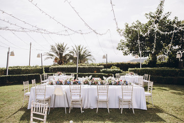 Wedding banquet. Tables in a green meadow are decorated with flower arrangements, on the tables are white tablecloths, plates with napkins, glasses and candles, cutlery. White bulbs hang over tables