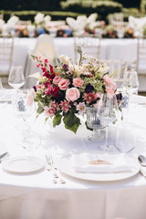 Wedding banquet. Tables in a green meadow are decorated with flower arrangements, on the tables are white tablecloths, plates with napkins, glasses and candles, cutlery. White bulbs hang over tables