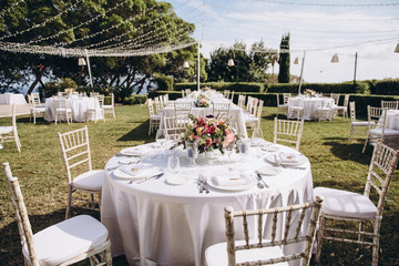 Wedding banquet. Tables in a green meadow are decorated with flower arrangements, on the tables are white tablecloths, plates with napkins, glasses and candles, cutlery. White bulbs hang over tables