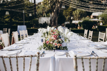 Wedding banquet. Tables in a green meadow are decorated with flower arrangements, on the tables are white tablecloths, plates with napkins, glasses and candles, cutlery. White bulbs hang over tables