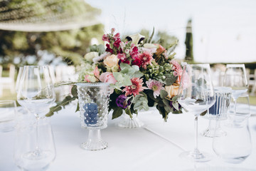 Wedding banquet. Tables in a green meadow are decorated with flower arrangements, on the tables are white tablecloths, plates with napkins, glasses and candles, cutlery. White bulbs hang over tables