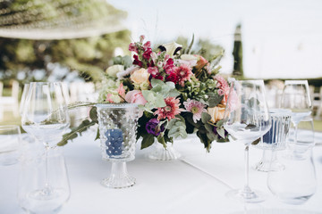 Fototapeta na wymiar Wedding banquet. Tables in a green meadow are decorated with flower arrangements, on the tables are white tablecloths, plates with napkins, glasses and candles, cutlery. White bulbs hang over tables