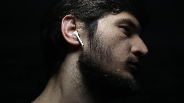 Dark close-up portrait of young man with wireless earphones on black background.