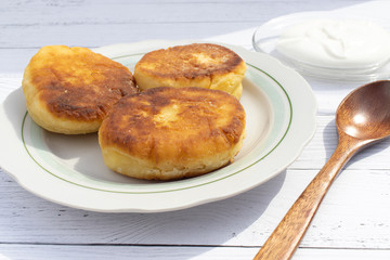 Three fried curd cheesecakes with a wooden spoon and sour cream on a transparent saucer on a white plate on a plank table. Delicious Breakfast of flour, eggs and cottage cheese