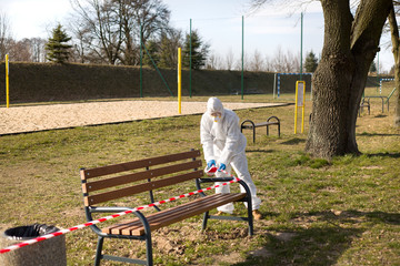 Osoba ubrana w kombinezon ochronny zabezpiecza ławki w parku by uniemożliwić korzystanie z nich w czasie pandemii - obrazy, fototapety, plakaty