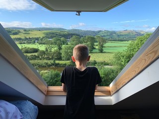 Boy looks out of a Velux window over a beautiful green valley on a summers morning