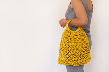 A girl in a gray dress stands with her back and holds in her hand an empty yellow reusable eco-friendly string bag.The concept of recycling,environmental care, naturalness.White background. Copy space
