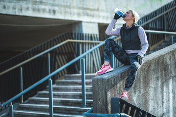 Young Caucasian woman taking a exercises break, sitting on the stair railing and drinking water 