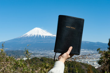 Raised hand holding the Holy Bible. Background with aerial view of Fuji City and Mount Fuji with...