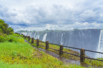Panorama view with dramatic clouds, walking way and waterfall at Victoria Falls on the Zambezi...