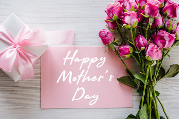 Happy mother's day text sign. Postcard with bouquet of small pink roses, gift box on white rustic wooden background. Greeting card with flowers concept. Holiday greeting card! Top view, flat lay.