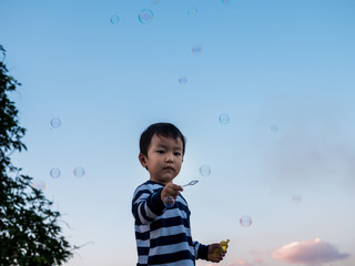 Asian child boy playing bubbles outdoor with happy face with nature blue sky background, bubbles in the air. Young kid have fun in relaxing holiday, freedom concept.