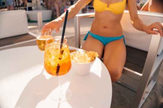 Picture of a girl sitting at the table in a beach cafe drinking beer with somebody wearing bikini, focus on first glass of spritz cocktail