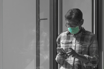 Asian man wearing protective mask and using smartphone near glass window in living room at home, black and white with splash green color techniques
