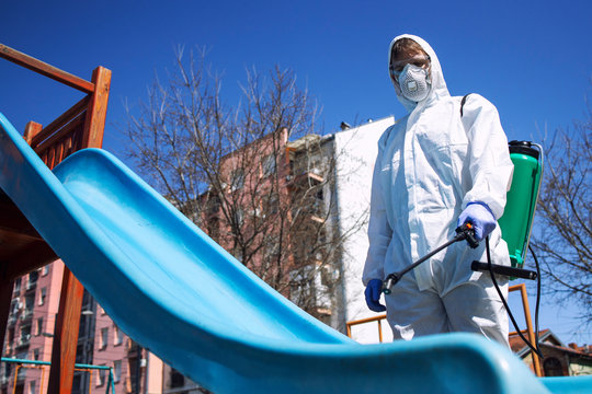 Children playground disinfecting and sanitizing against virus and disease. Man in white protective suit spraying disinfectant on slide to stop spreading coronavirus or COVID-19.