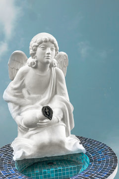 White statue of an angel by the bowl of holy water on his knees with a jug in his hands and an Orthodox cross isolated against a blue sky.