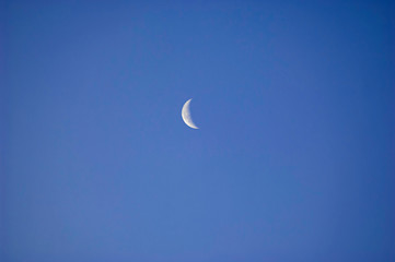 A slim sliver of the silvery moon set on an expanse of clear and cloudless blue sky.