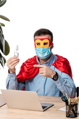 man in medical mask and superhero cloak pointing with finger at hand sanitizer near laptop isolated on white