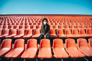 Curly girl in a surgical mask sitting in an empty stadium during epidemic disease Covid-19.