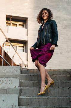 Young european female with short curly hairy in a classy long magenta dress black jacket and yellow heels walking down stairs laughing