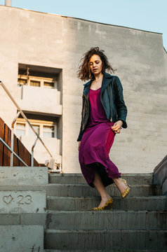 Young european female with short curly hairy in a classy long magenta dress black jacket and yellow heels walking down stairs