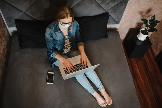 Coronavirus. Business woman working in home, wearing protective mask in quarantine. Stay at home. Girl learns, using laptop computer in bedroom. Freelancer. Communication and technology concept.