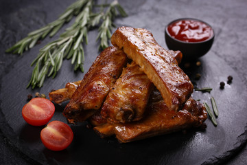Fried pork ribs with a fried crust with spices, rosemary, cherry tomatoes and ketchup in bowl on a black board on a black background. Meat on grill. Background image, copy space