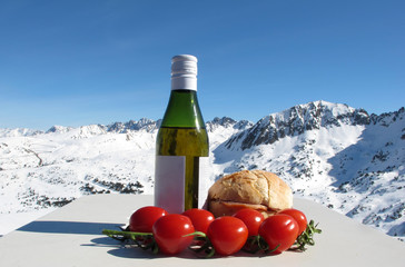 A small bottle of wine, a homemade sandwich and cherry tomatoes against a background of snow-covered mountains and a bright blue sky. Travel content