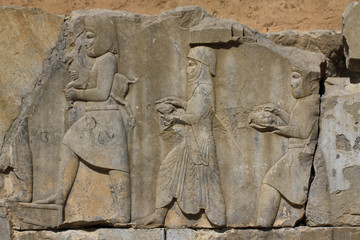 starożytne płaskorzeźby wykute w blokach kamiennych przedstawiające  mężczyzn idących z darami na jednej ze ścian w persepolis w iranie - obrazy, fototapety, plakaty