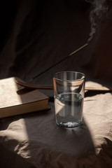 The composition is a glass cup half filled with water, a book and a smoke stick on a cloth background. Soft light. Trends color