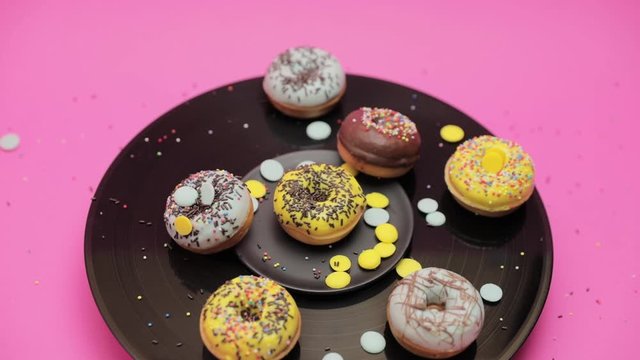 A bunch of American donuts with black, yallow and blue chocolate and black and multicolored crumbs rotate on pink and black backgrounds in a circle