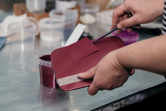The painter’s hands hold a test plate with a paint applied on it, matched to the color of the car’s hatch and the burgundy car part