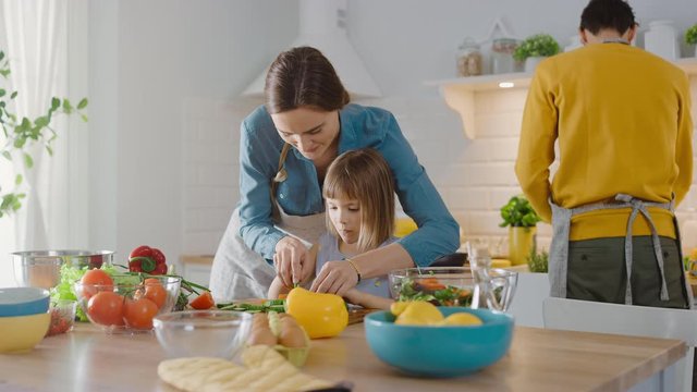 In Kitchen: Mother and Cute Little Daughter Cooking Together Healthy Dinner. Mom Teaches Little Girl Healthy Habits and how to Cut Vegetables for Salad. Cute Child Helping Her Beautiful Caring Parents