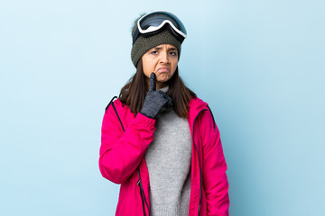 Mixed race skier girl with snowboarding glasses over isolated blue background having doubts while looking up.