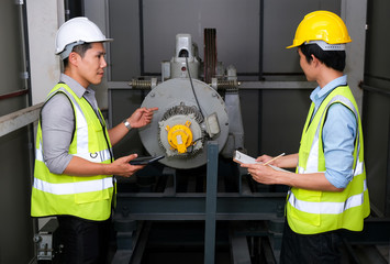 Senior engineer point to the lift motor and explain to junior engineer. Feeling fun on work at the control room of plant. Wearing safety vest and safety helmet. Engineering and control room concept.