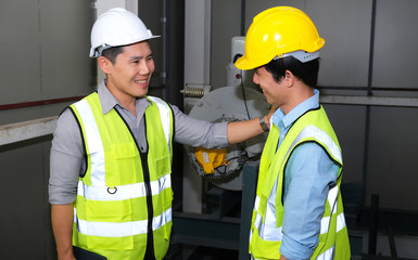 Senior engineer touch the shoulder of junior engineer. They  feeling happy and fun on finishing work at the control room of plant. Wearing safety vest and helmet. Engineering and control room concept.