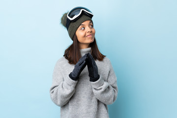 Mixed race skier girl with snowboarding glasses over isolated blue background scheming something.